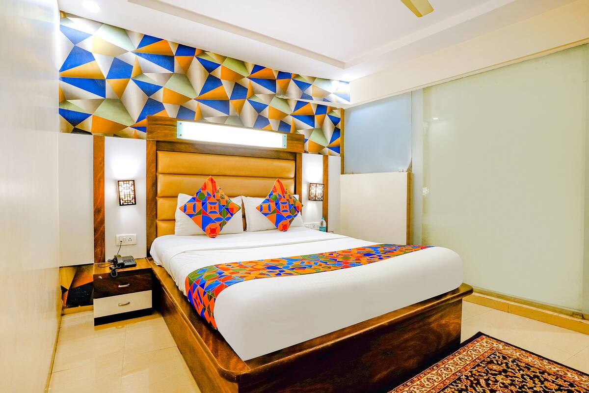 A modern hotel room with a geometric-patterned headboard, a neatly made bed with colorful pillows, a bedside table with a lamp, and a large window.