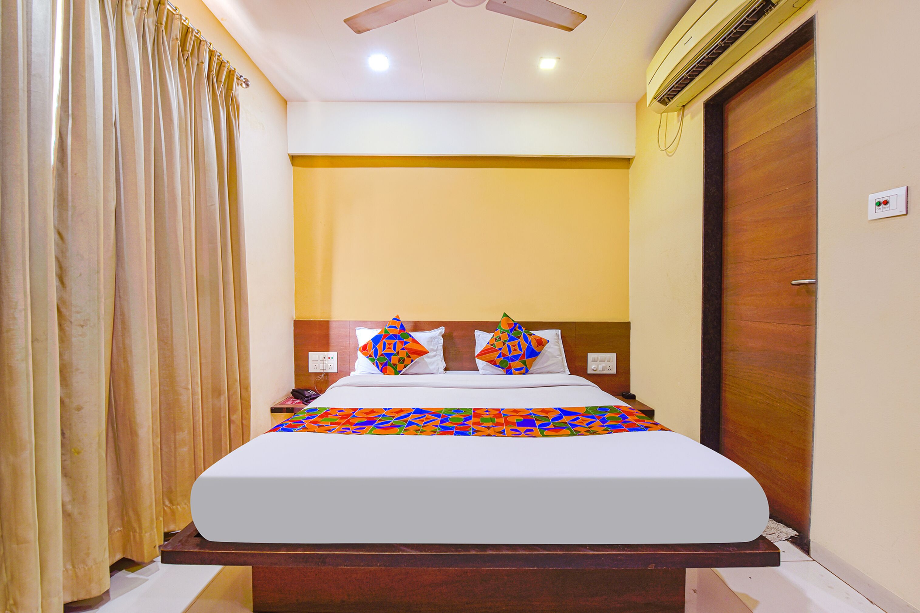 A hotel room with a large bed, a wooden headboard, a ceiling fan, an air conditioning unit, and a wooden door.