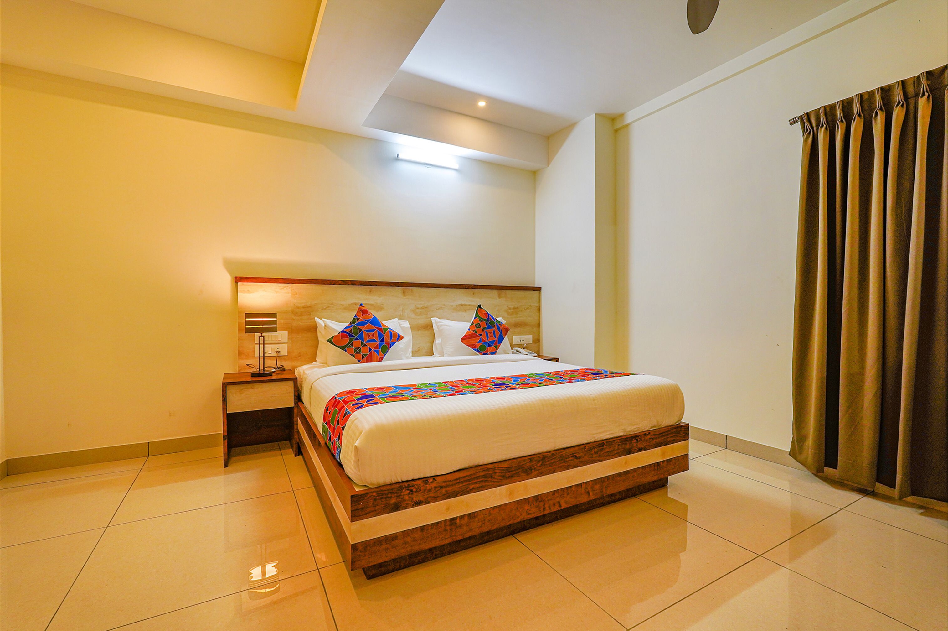 A neatly made bed with a wooden frame and a colorful quilt, a bedside table with a lamp, and a window with curtains.