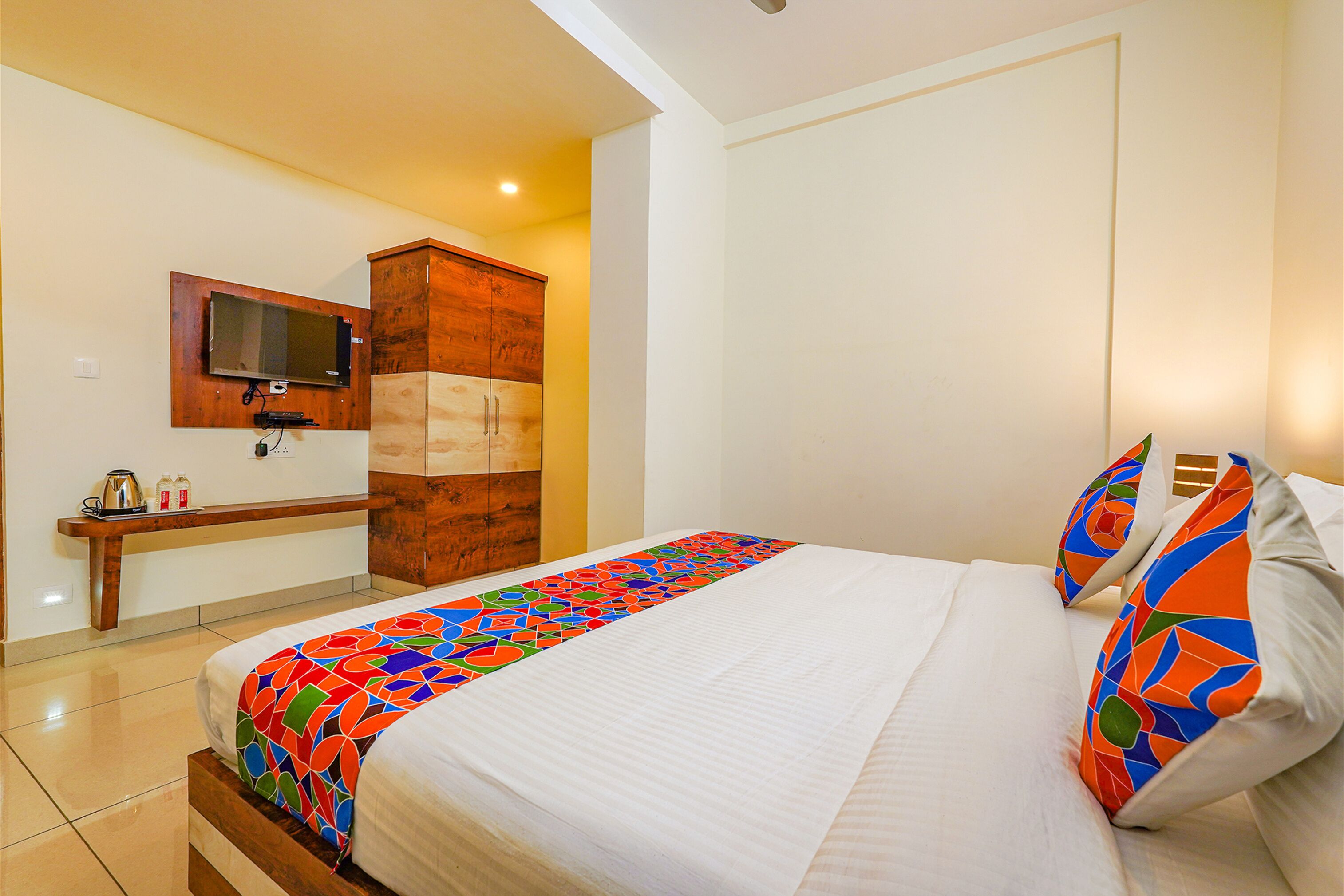 A hotel room with a bed, a TV mounted on the wall, a wooden cabinet, and a small table with a kettle and bottles.