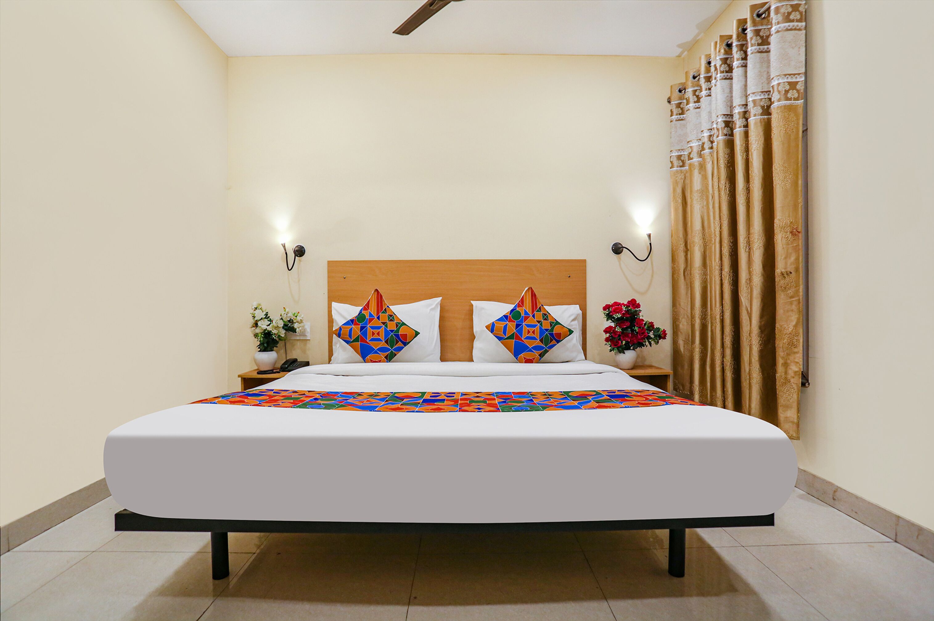 A neatly made bed with a white mattress and colorful geometric pillows, a wooden headboard, and a nightstand with a vase of flowers.
