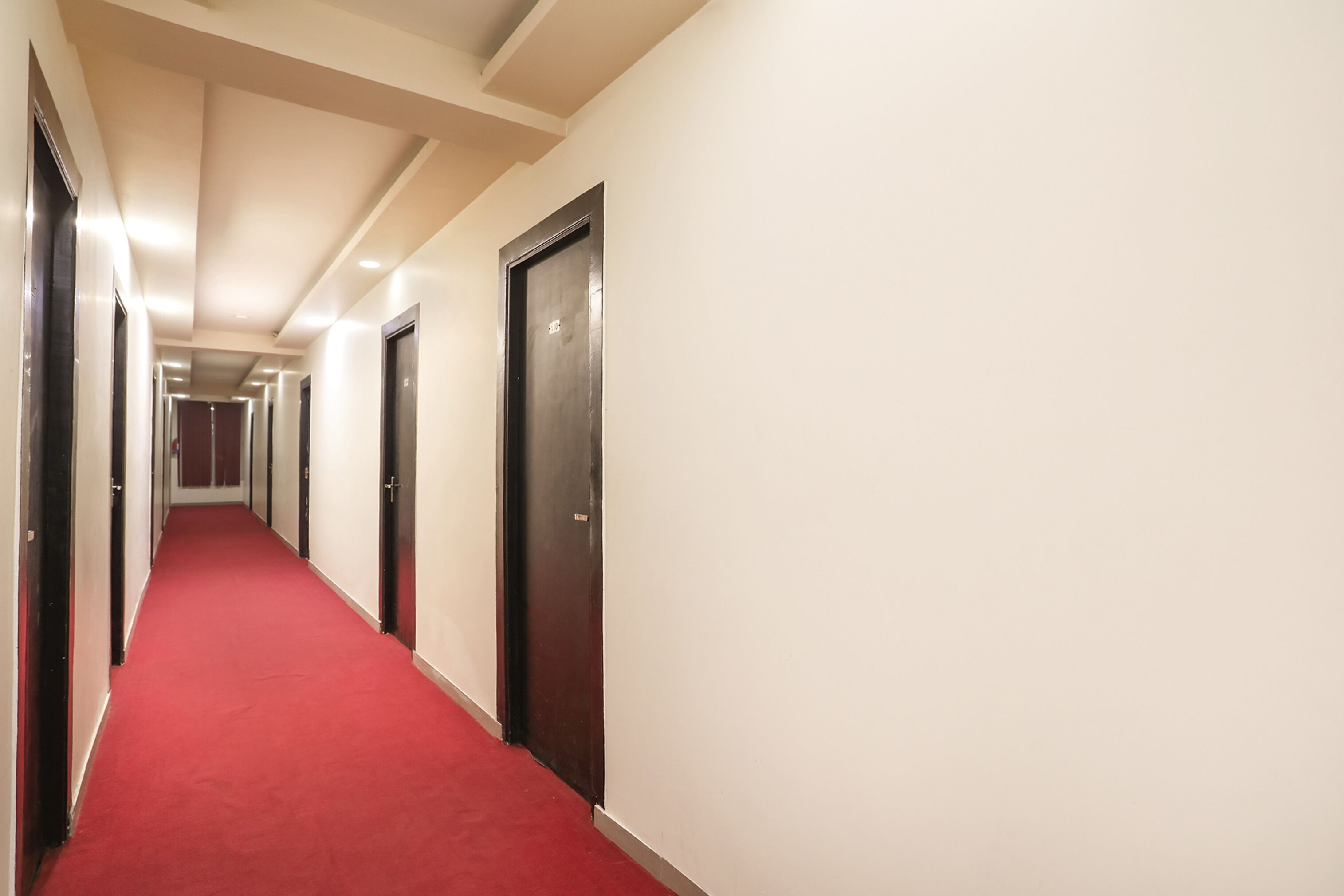A long, narrow hallway with red carpet and white walls, featuring doors on both sides and a ceiling with recessed lighting.