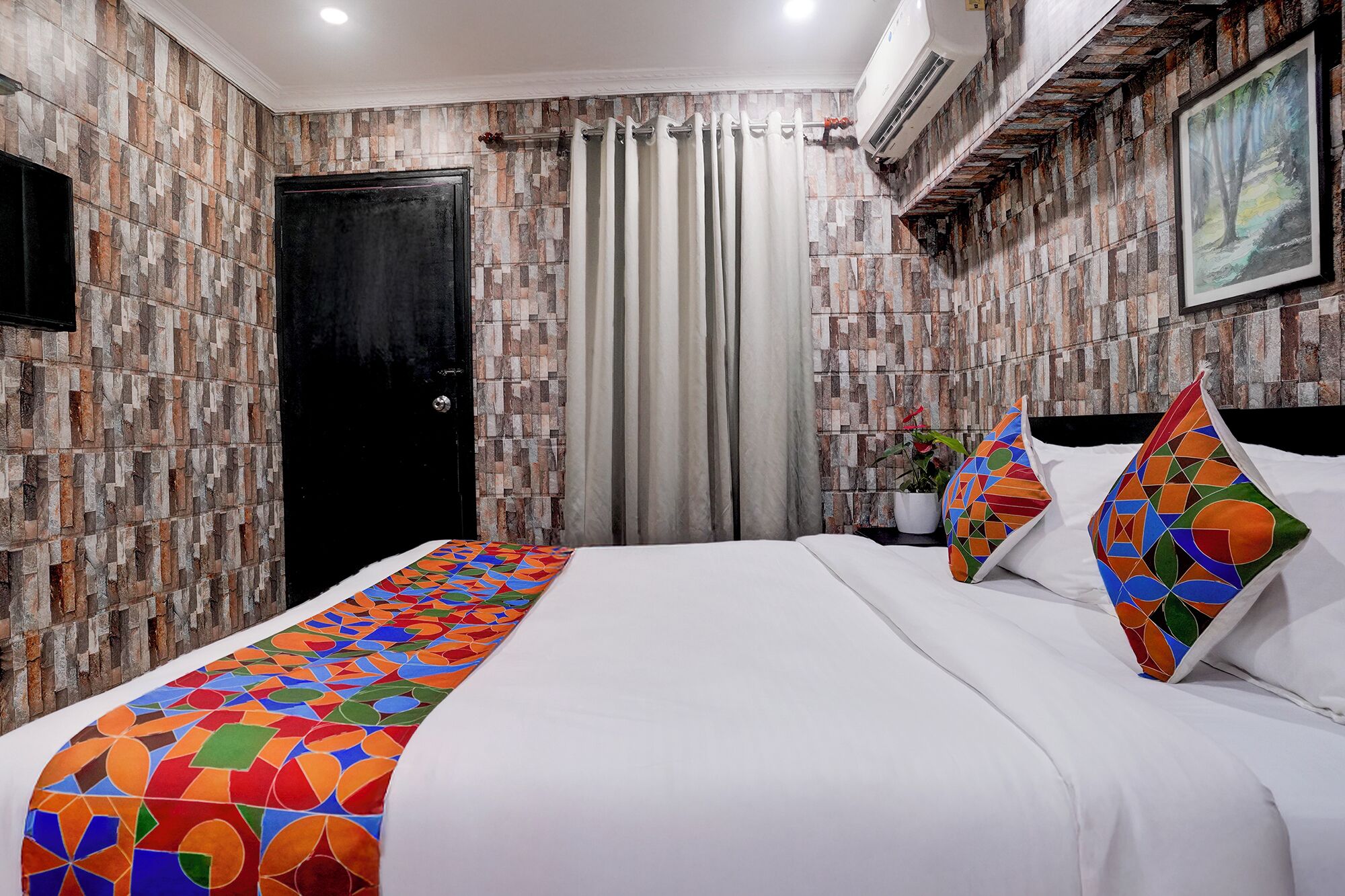A hotel room with a bed, a black door, a curtain, a potted plant, and a painting on the wall.
