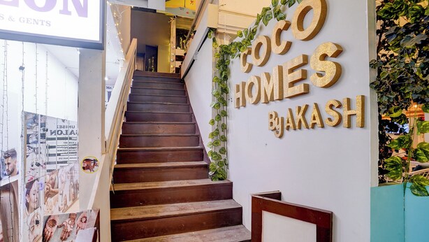 Un escalier en bois mène à un palier. Des affiches sont accrochées aux murs, et une enseigne indique : « Coco Homes By Akash ».
