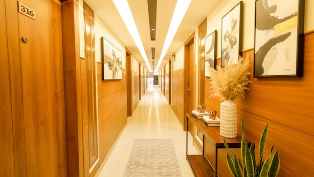 Un pasillo de hotel con paneles de madera, puertas numeradas, obras de arte en las paredes y un mostrador de recepción con una planta.