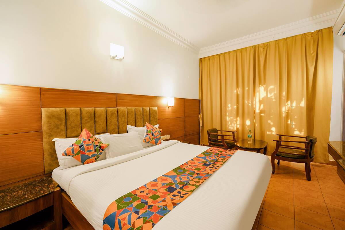 A hotel room with a large bed, a wooden headboard, a bedside table, a chair, a small table, and a curtain.