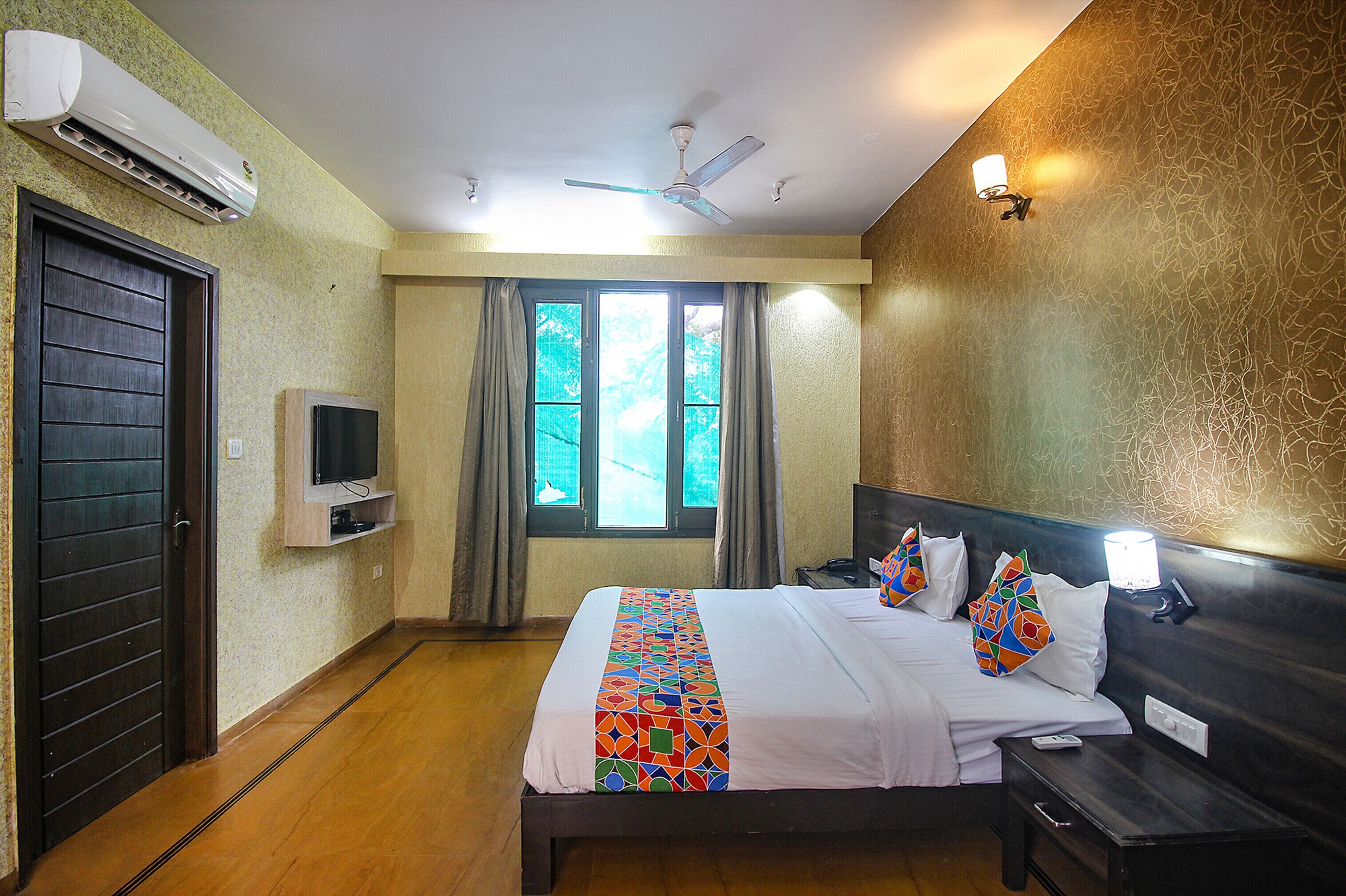 A hotel room with a bed, a TV, a ceiling fan, a window with curtains, and a wall-mounted light.