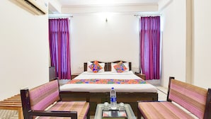 Une chambre d’hôtel avec un lit, deux chaises, une petite table avec une bouteille d’eau et une fenêtre ornée de rideaux violets.
