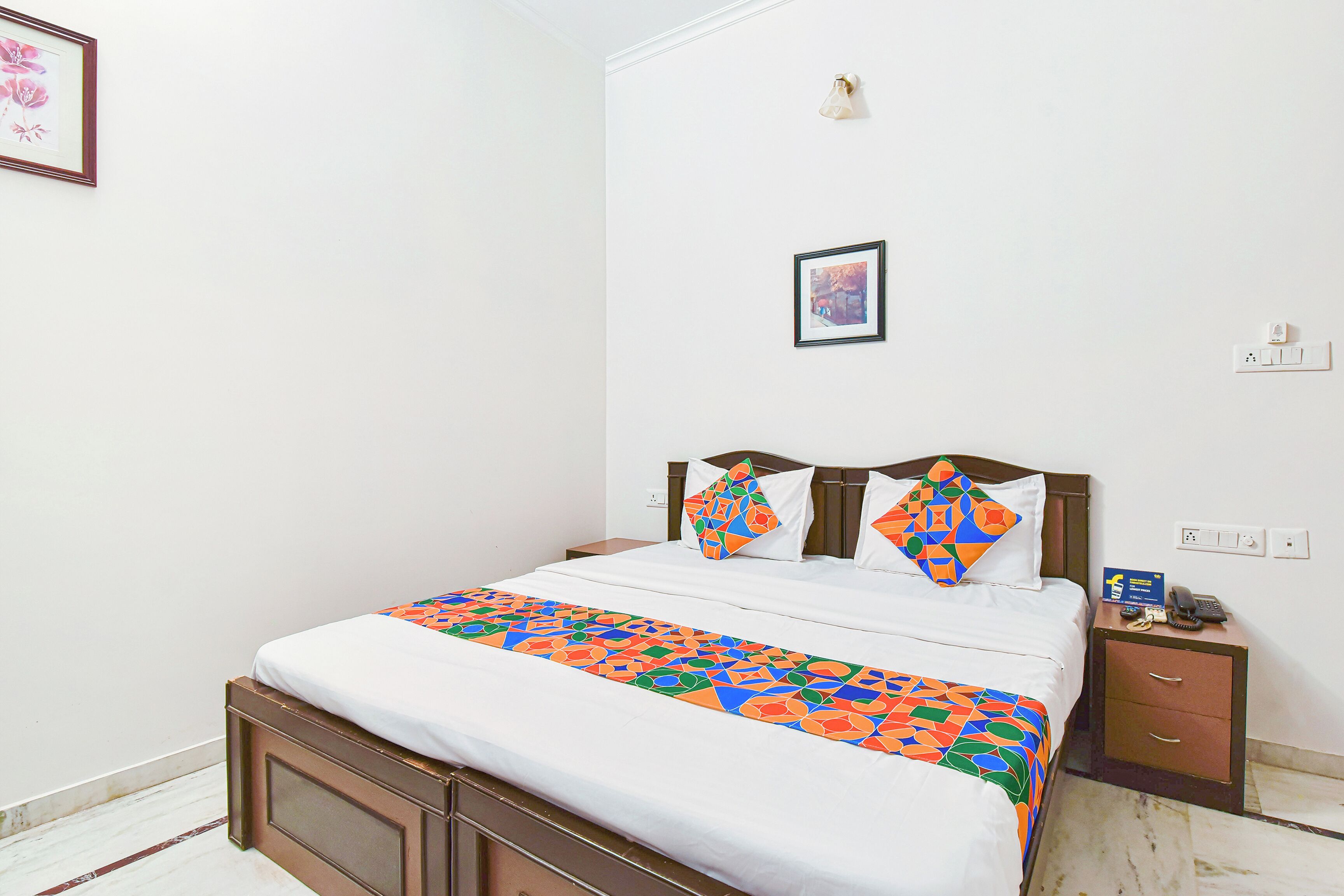 A neatly made bed with a colorful bedspread, a bedside table with a phone and a small box, and a framed picture on the wall.