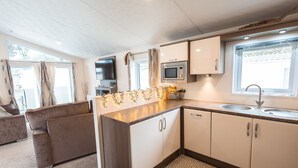 A compact kitchen with a microwave, sink, and cabinets. There is a sofa and a window with curtains.