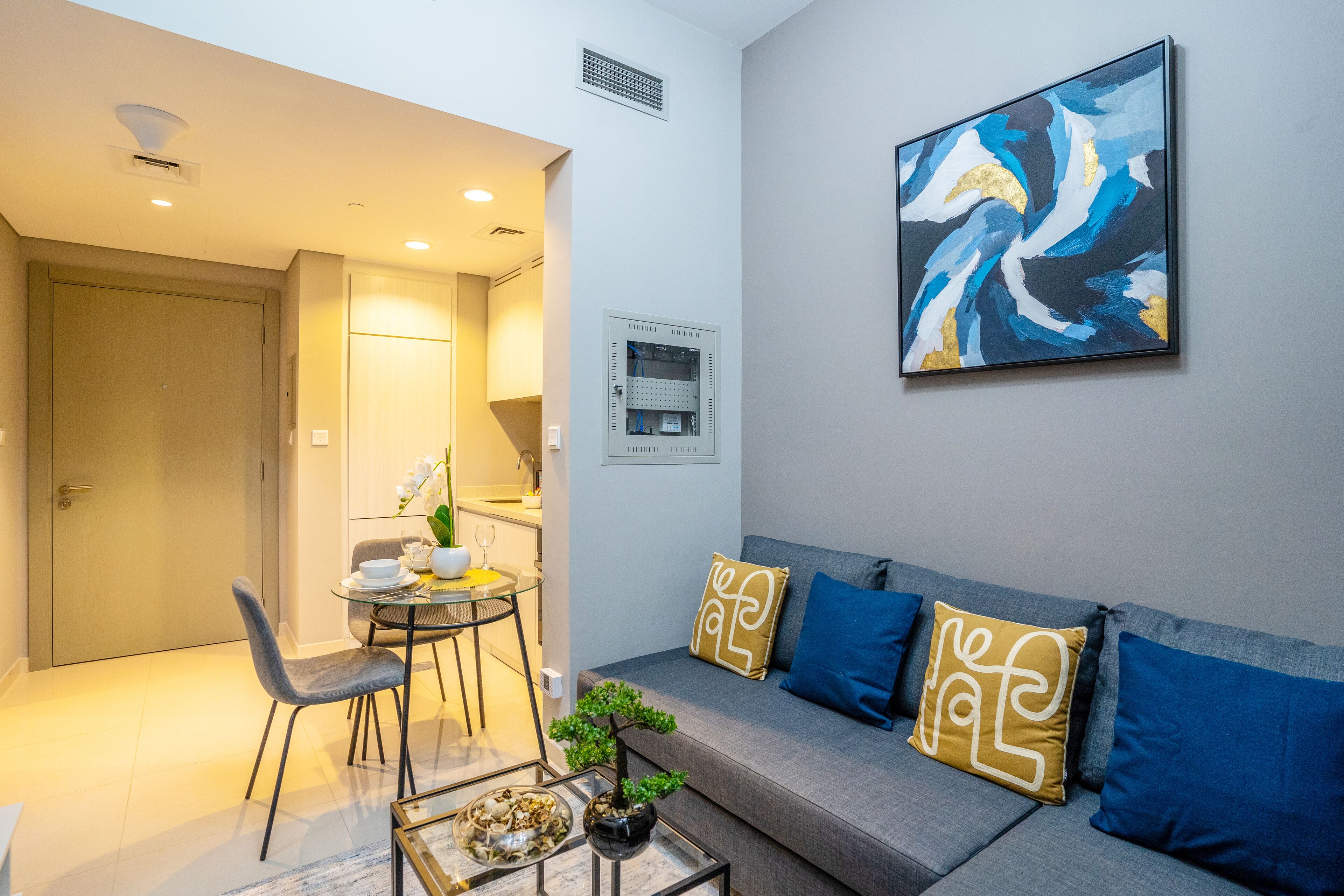 A modern living room with a grey sofa, blue and gold cushions, a glass coffee table, and a small plant.