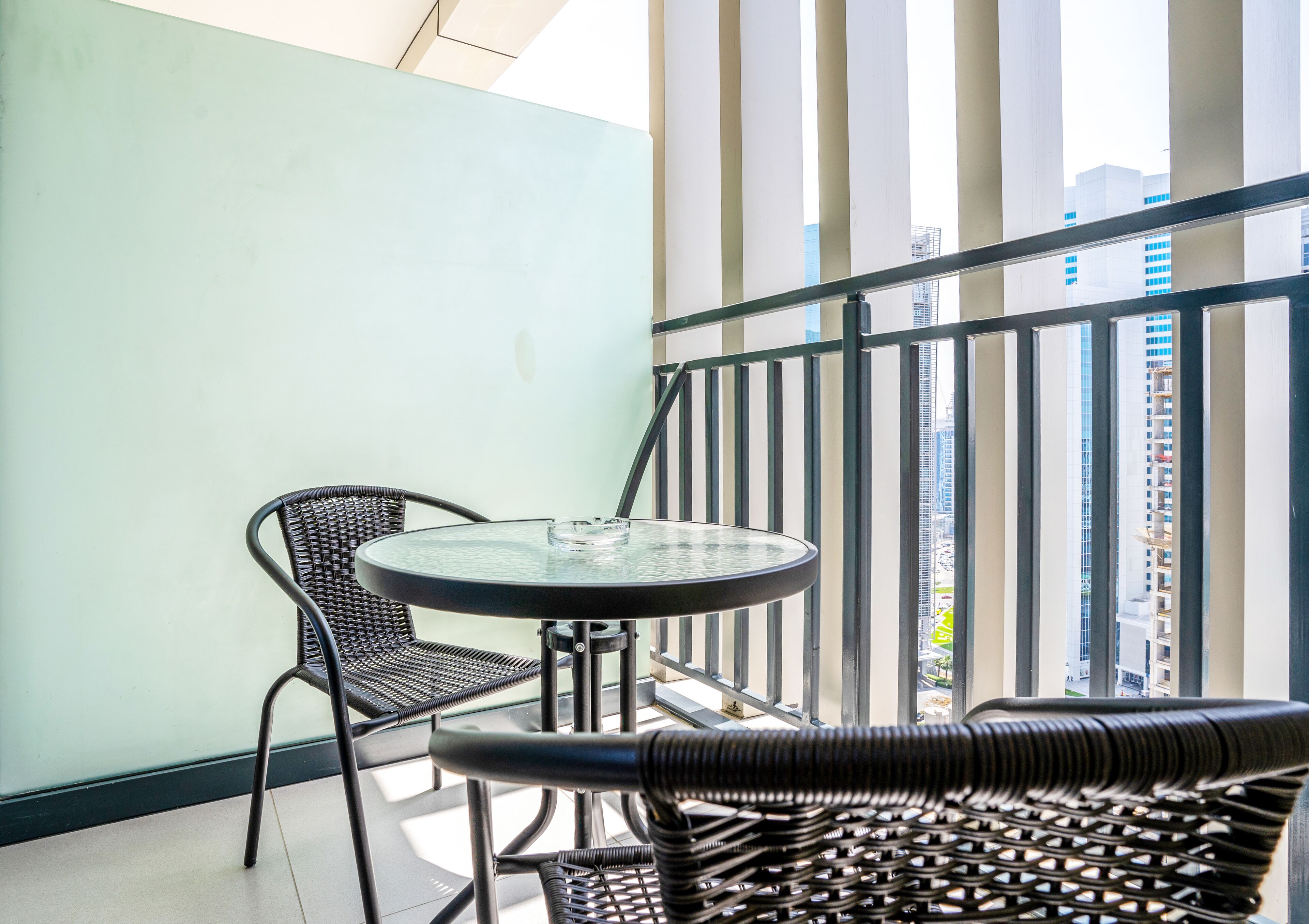 A balcony with a round glass table and two wicker chairs, overlooking a cityscape.