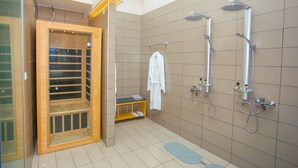 Un baño moderno con sauna de vidrio, banca de madera, toalla blanca colgada y ducha con varios cabezales.