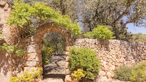 Un arco de piedra conduce a un camino, flanqueado por macetas con plantas y un muro de piedra.