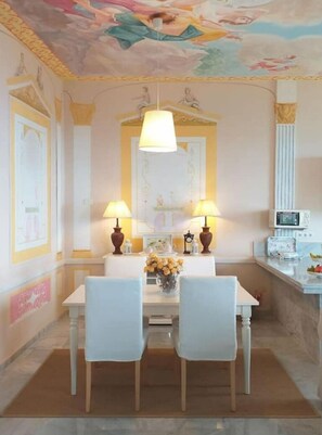 A dining area with a table for two, flanked by two chairs, features a pendant light above and a colorful mural on the ceiling.