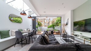 A modern living room with a large black sofa, dining area with chairs, and a TV mounted on the wall. - 5 Bed Apartment in Clapham (London)