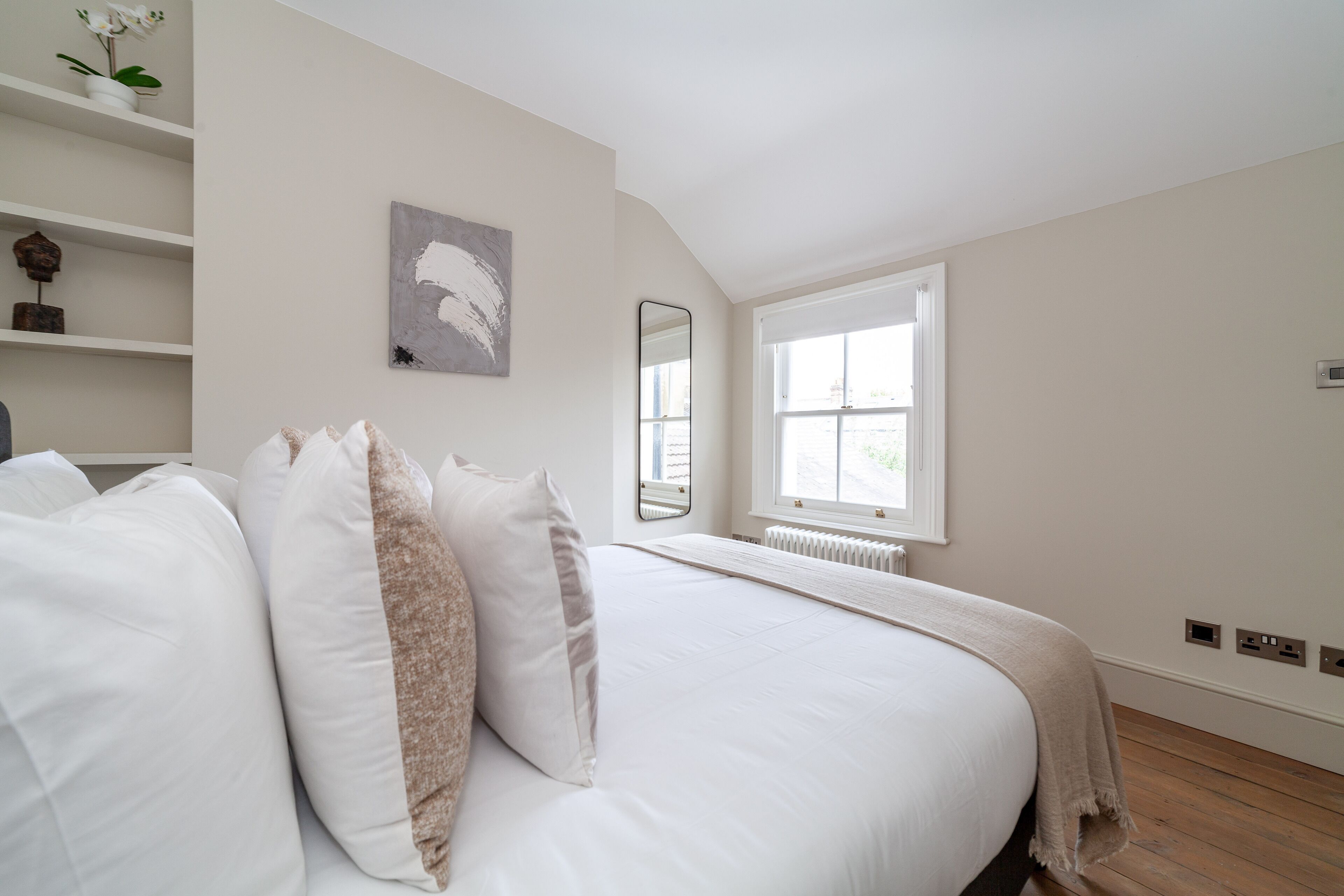 Een slaapkamer met een groot bed, wit beddengoed, een raam met uitzicht, een radiator en een plank met decoratieve voorwerpen.