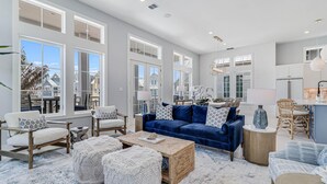 A spacious living room with a blue sofa, white armchairs, and a coffee table. Large windows provide ample natural light.