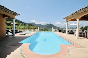 Una piscina rectangular con un emblema decorativo en el centro, rodeada de sillas de descanso y sombrillas, y un paisaje montañoso al fondo.