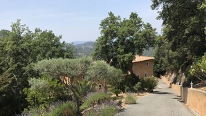 A narrow paved road flanked by dense greenery and a building with a red-tiled roof. - Bastide Provençale au Cœur du Massif de L'esterel (FREJUS)