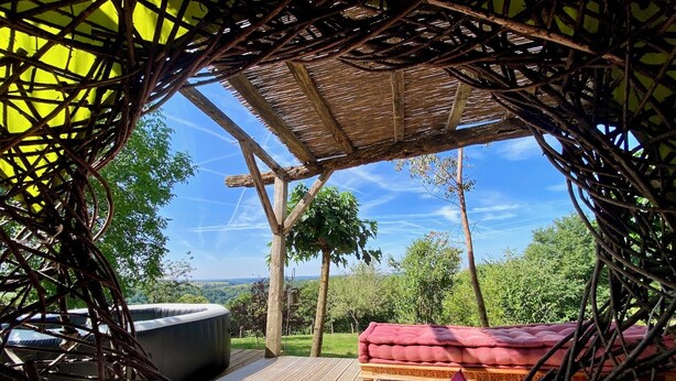 An outdoor seating area with a woven canopy, a cushioned bench, and a round table. - Eco Gite Les Monts D'amara (CAMPUAC)