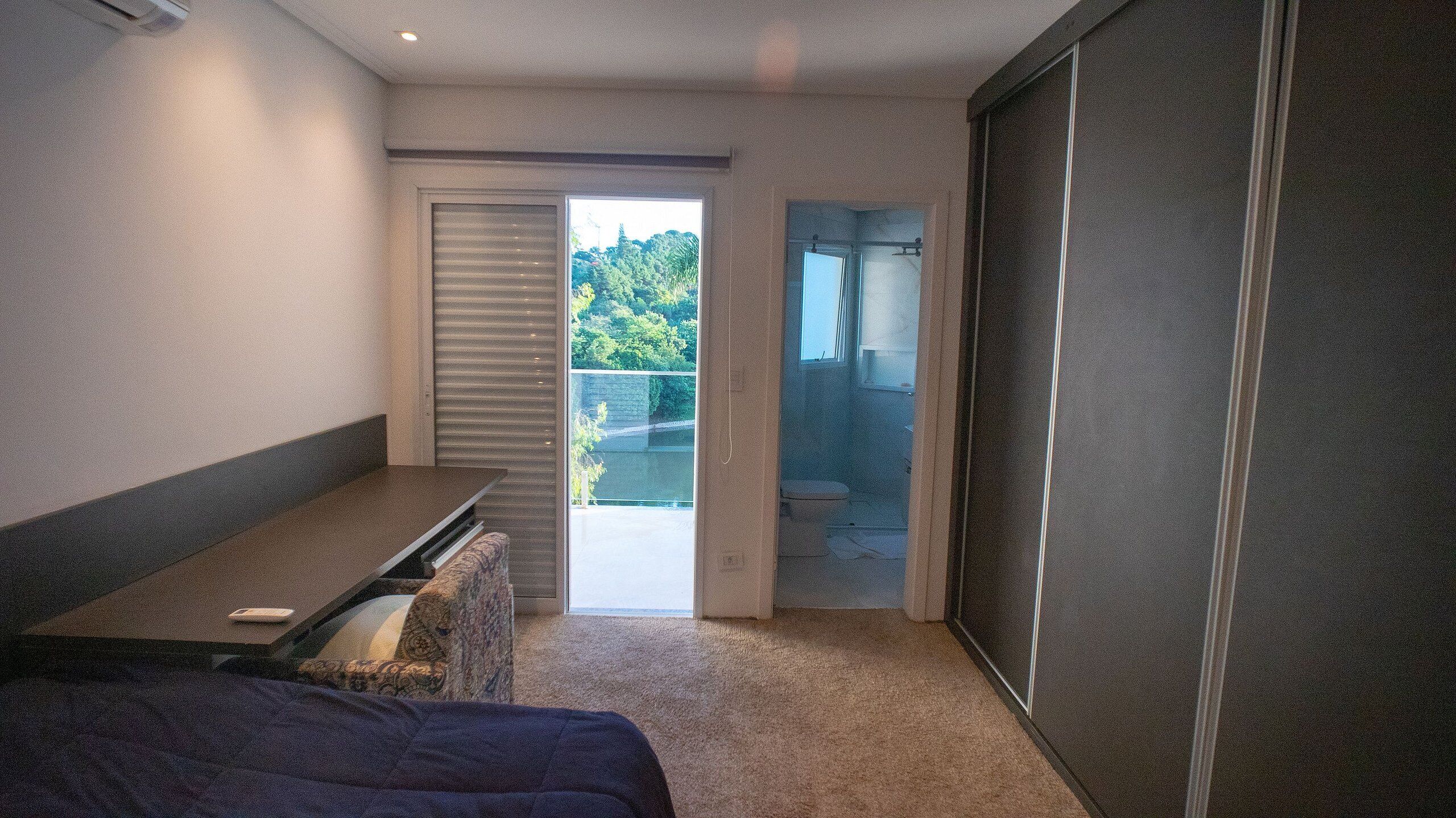 A bedroom with a bed, a desk, and a view of a balcony with a door leading outside.