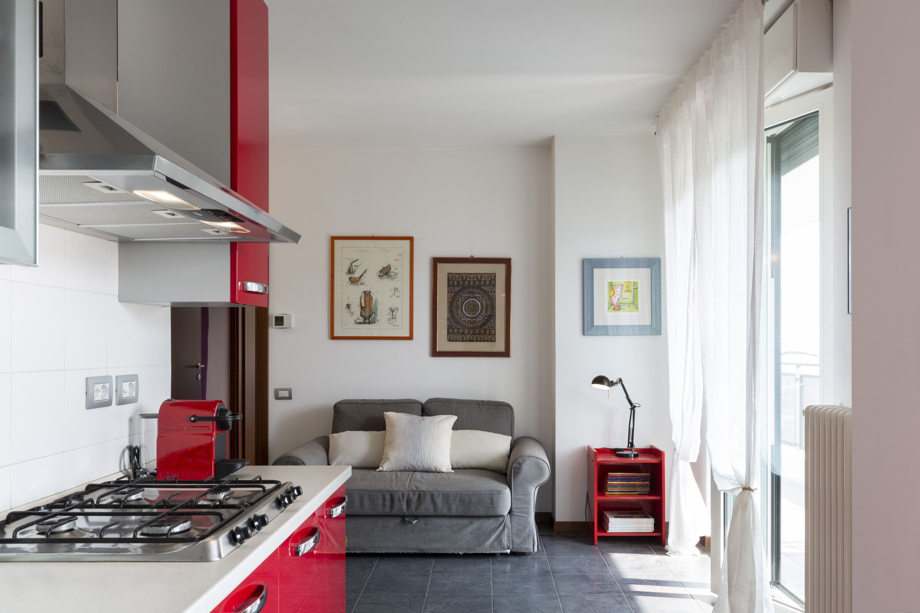 Cocina compacta con estufa a gas, electrodoméstico rojo y sofá gris. En la pared hay cuadros enmarcados y una ventana con cortinas.