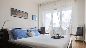 Habitació d'hotel moderna amb un llit gran, una taula de treball, una cadira, un radiador i una finestra amb cortines fines.