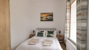 Ein ordentlich bezogenes Bett mit Kissen und gefalteten Handtüchern, eine Nachttischlampe und ein Sonnenuntergangsbild an der Wand.