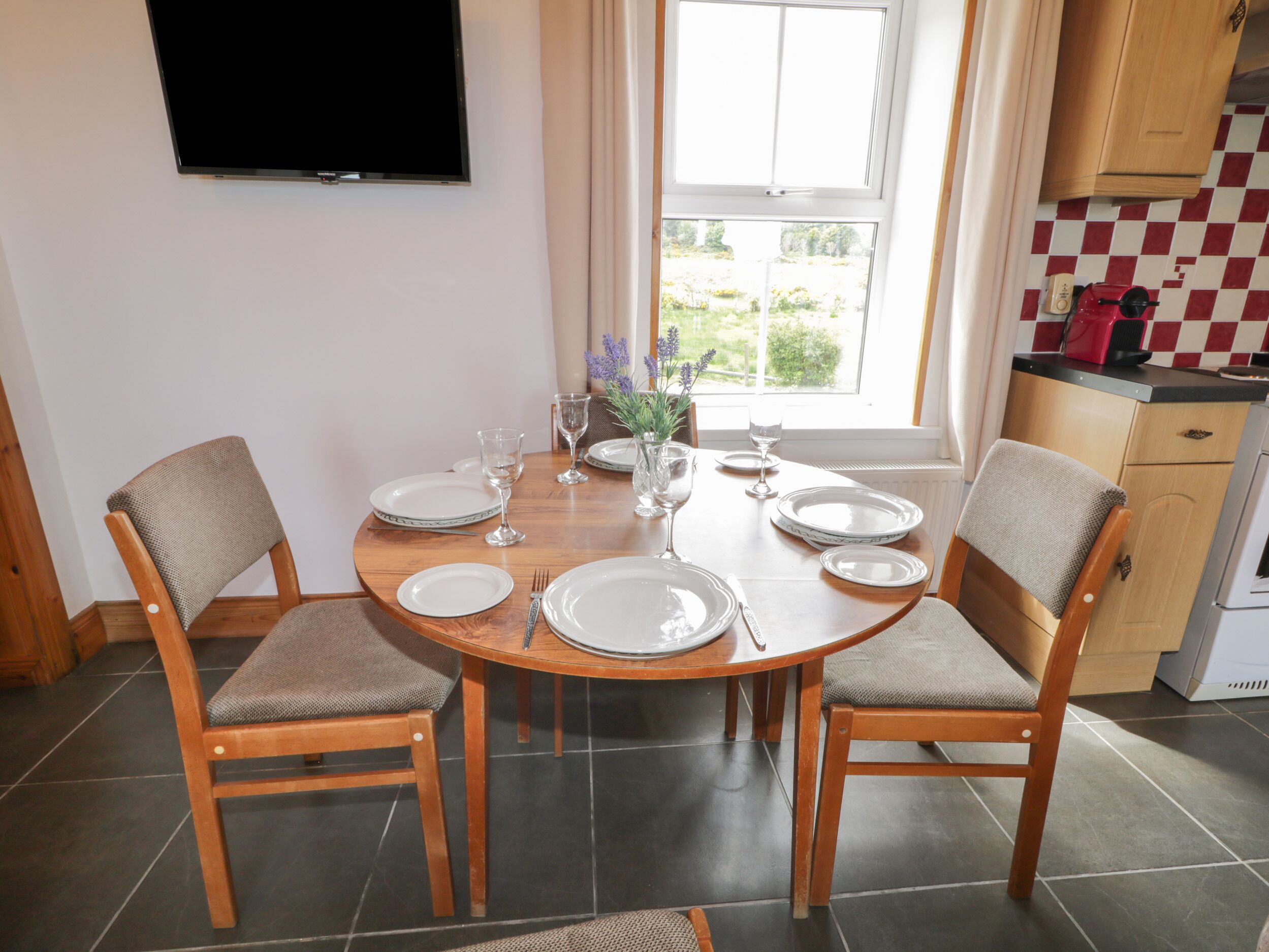 A dining area with a round wooden table set for four, surrounded by chairs with cushioned seats. A flat-screen TV is mounted on the wall above the table.