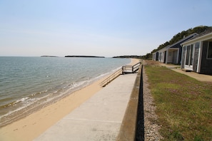 A beachfront property with a concrete walkway leading to a sandy beach. There are several small buildings with large windows along the shore.