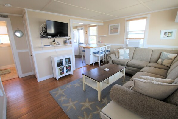 A compact living space with a sofa, a coffee table, a TV mounted on a cabinet, a small kitchen area, and a dining table.