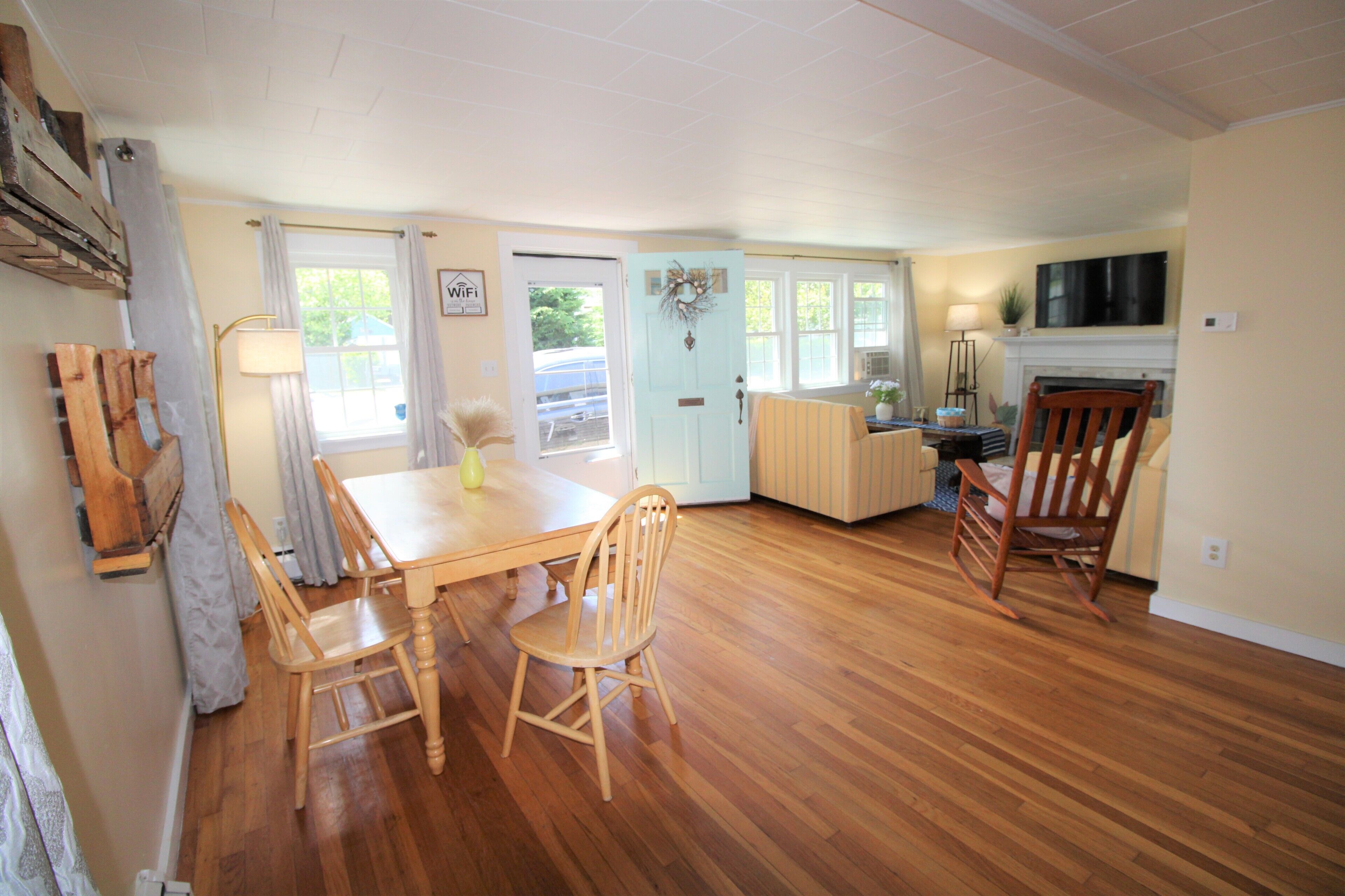 A spacious living room with a wooden floor, a dining table with chairs, a fireplace, and a television mounted on the wall.