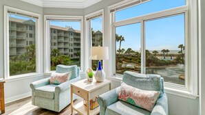Habitación de esquina con dos sillones, una mesita y ventanas amplias con vistas a un edificio y una playa.