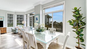 Un amplio comedor con una mesa de vidrio y sillas blancas, ventanas grandes que ofrecen vistas al exterior y un ventilador de techo.
