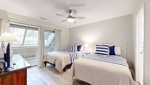 Une chambre avec deux lits, un ventilateur de plafond, un bureau avec une lampe et une vue sur l’extérieur grâce à de grandes fenêtres.