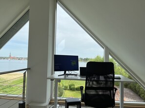 Un espace de travail équipé d’un ordinateur, d’une chaise et d’un bureau, situé près d’une grande fenêtre donnant sur une rivière et des bâtiments éloignés.