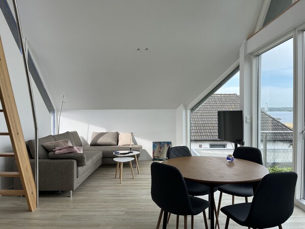 Un salon moderne avec un canapé gris, une table ronde en bois entourée de chaises et une grande fenêtre donnant sur un paysage urbain et l’eau.