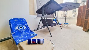 Un balcón con una silla azul estampada, una silla lounge negra y una pequeña mesa con cojín a rayas.
