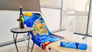 Un balcón con una silla tapizada con un mapa de colores, una mesita con una botella de vino y una toalla enrollada.