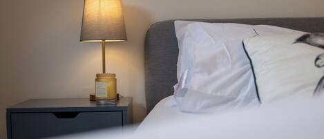 A neatly made bed with white linens, a gray headboard, and a bedside table with a lamp and a candle.