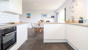 A modern kitchen with white cabinetry, a built-in oven, and a dining area with a table and chairs. - Charlotte's House (Penzance)