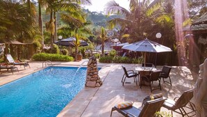 A swimming pool with lounge chairs and umbrellas, surrounded by palm trees and a stone structure. - Pousada Villa 3 Lagos (Santo Antonio do Pinhal)