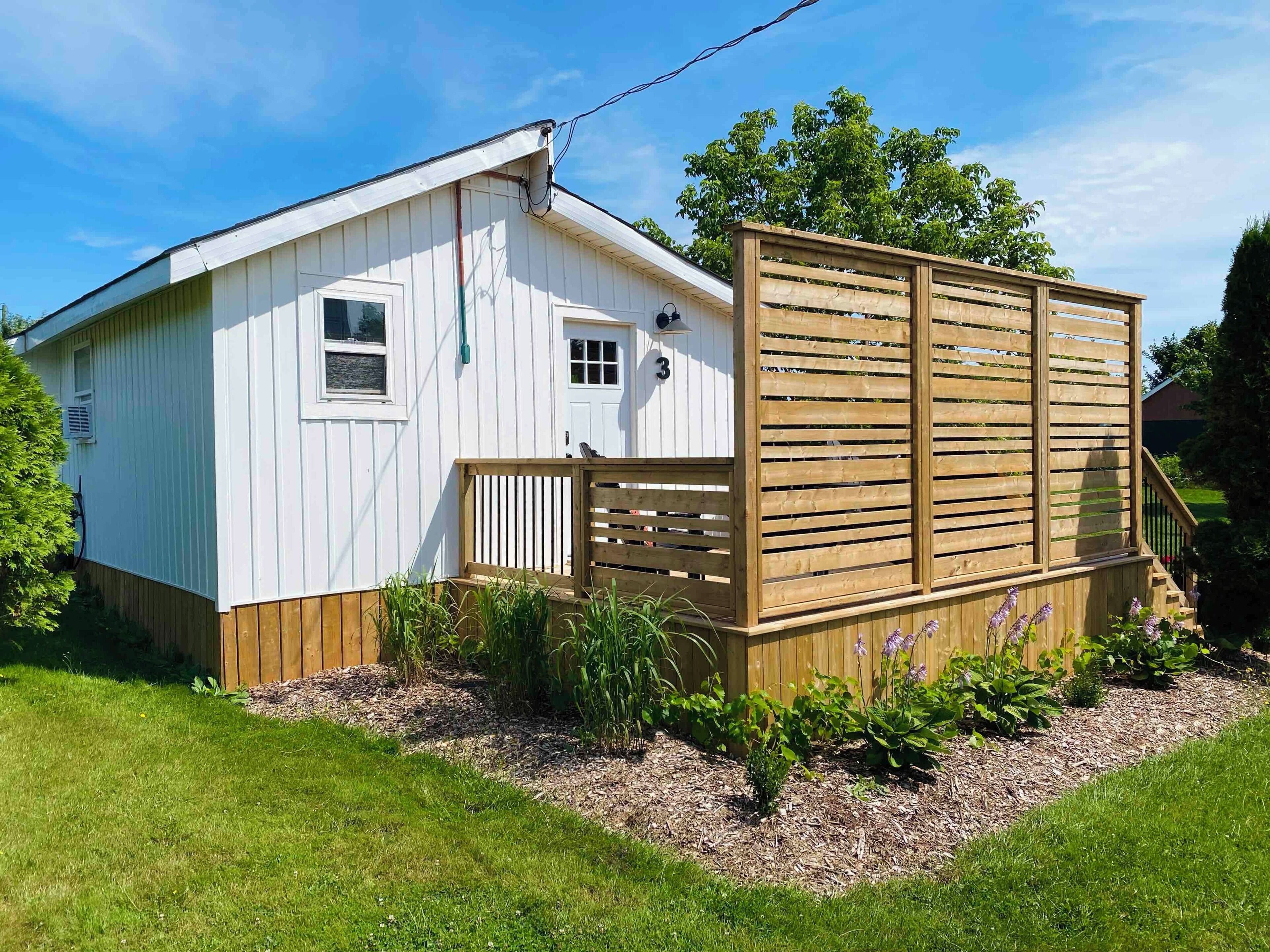 A single-story white building with a wooden deck and a fenced area with plants.
