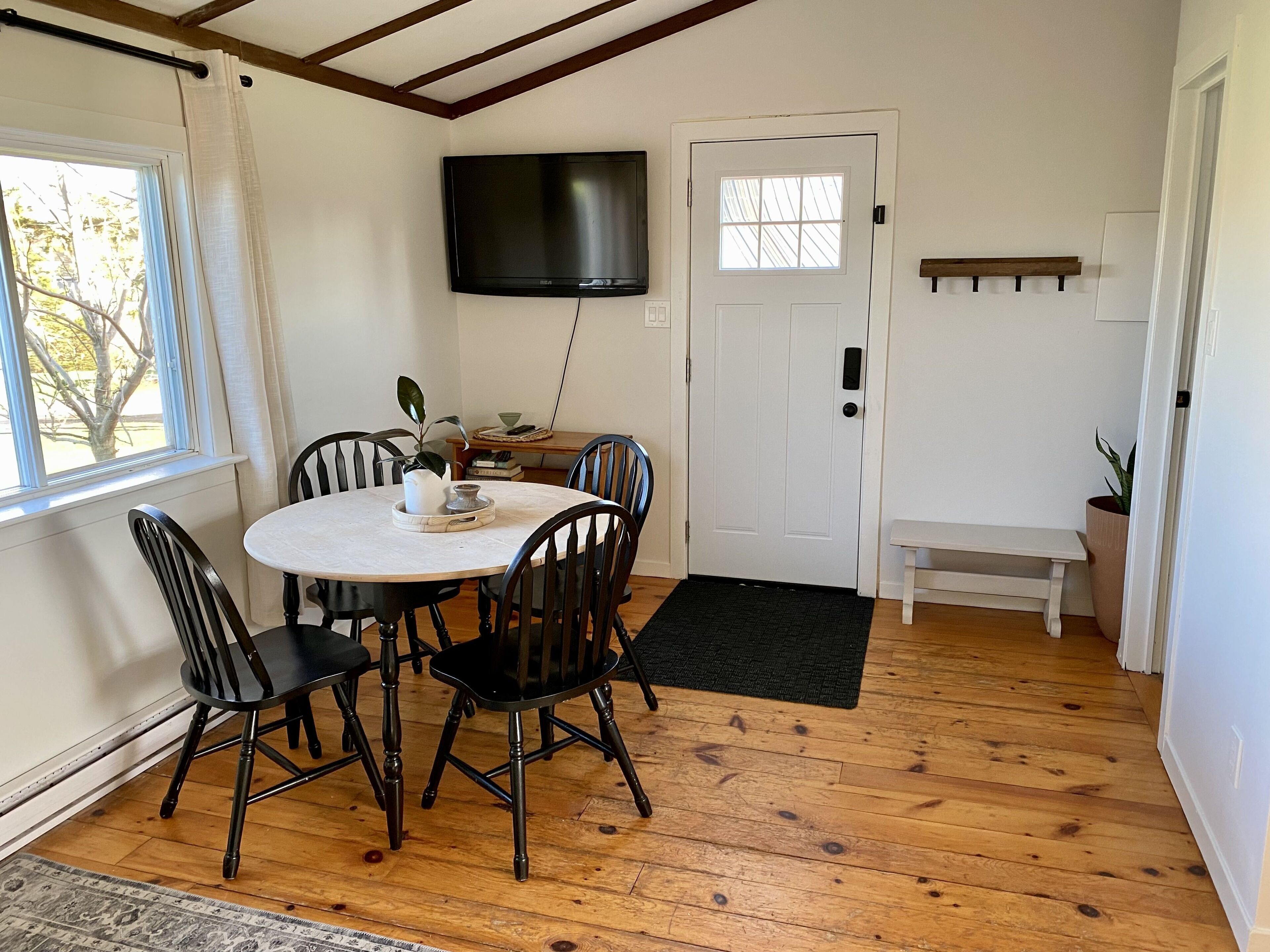 A dining area with a round table, four chairs, a TV mounted on the wall, a white door, and a bench.