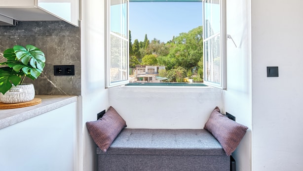 Habitación con un banco empotrado, una planta en maceta y vistas de una casa a través de una ventana abierta.