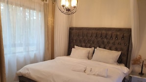 Una cama bien tendida con un cabecero capitonado, una mesita de noche con cajones y cortinas traslúcidas en la ventana.