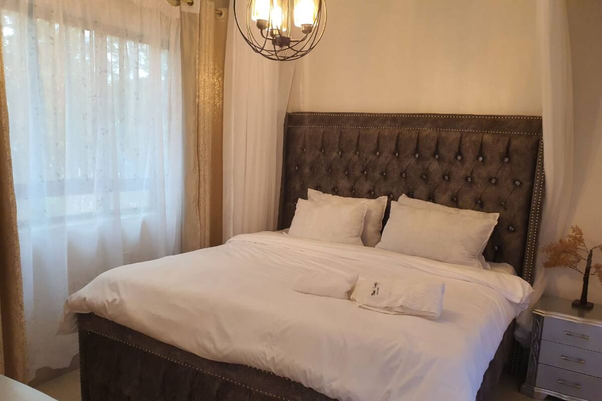 A neatly made bed with a tufted headboard, a bedside table with drawers, and sheer curtains on a window.