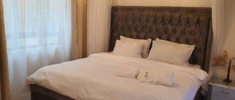 A neatly made bed with a tufted headboard, a bedside table with drawers, and sheer curtains on a window.