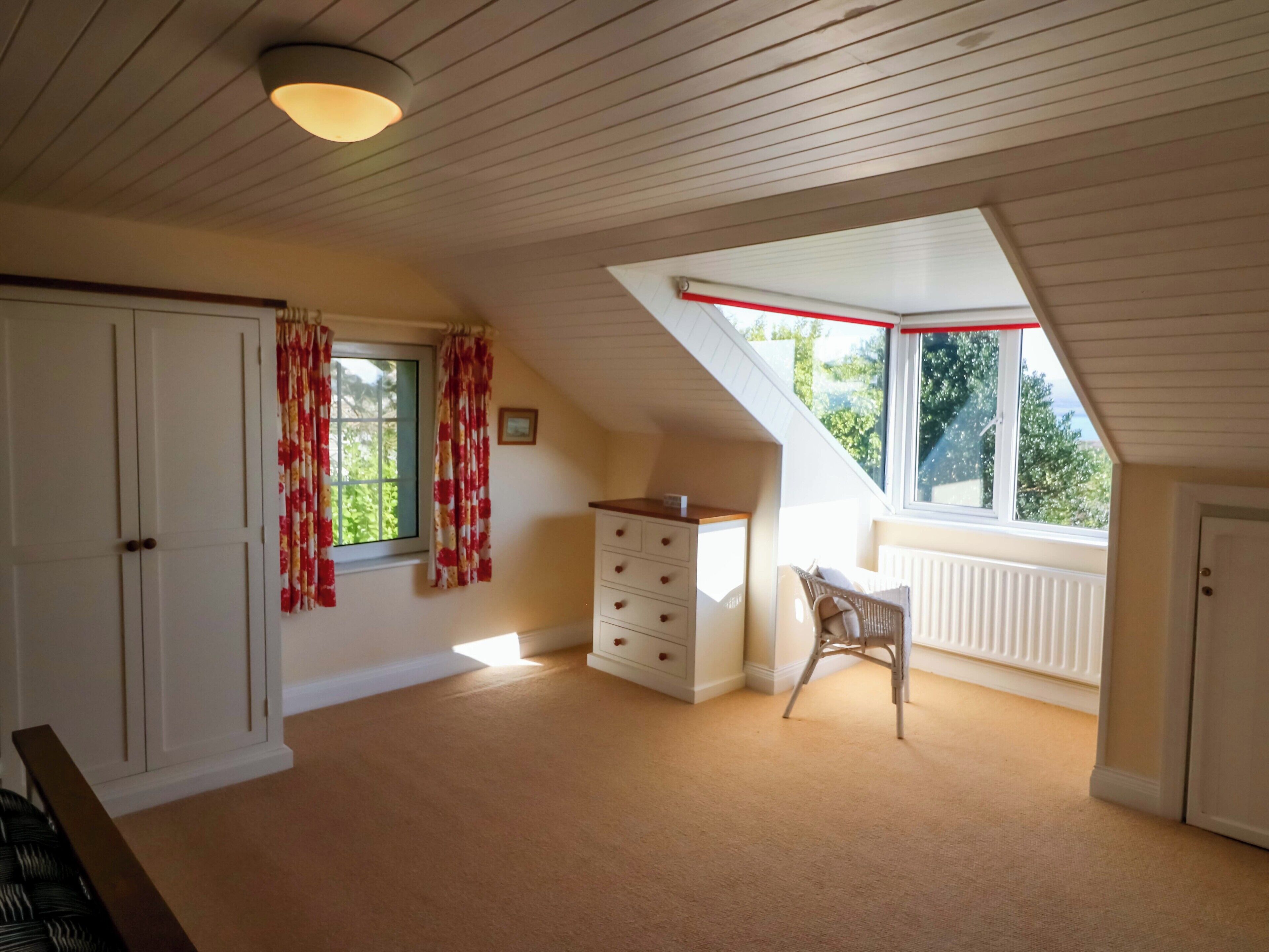 A room with a sloped ceiling, a window with a red trim, a white dresser, a chair, and a radiator.